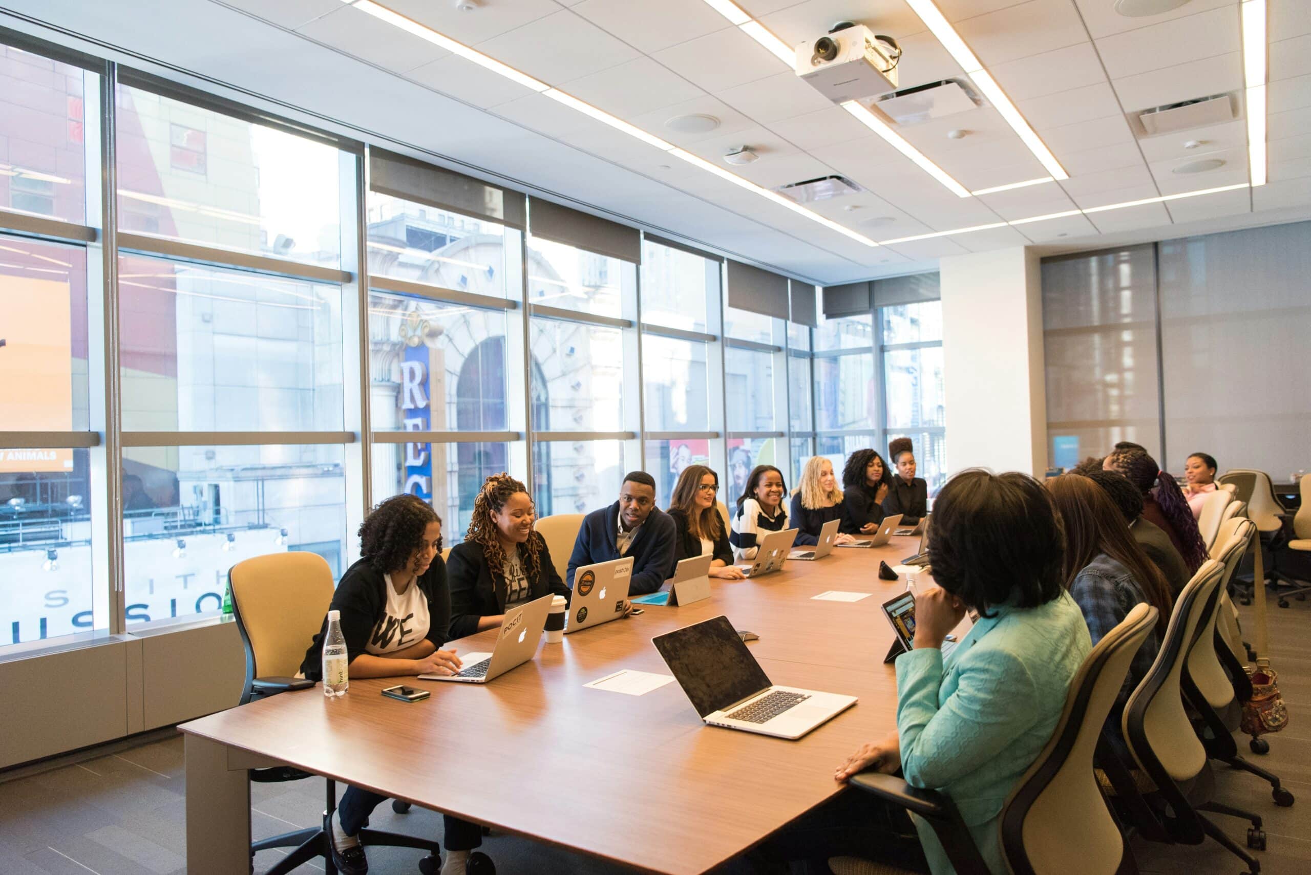 A team of business professionals seated at a long conference table chatting and working on their laptops and notebooks.