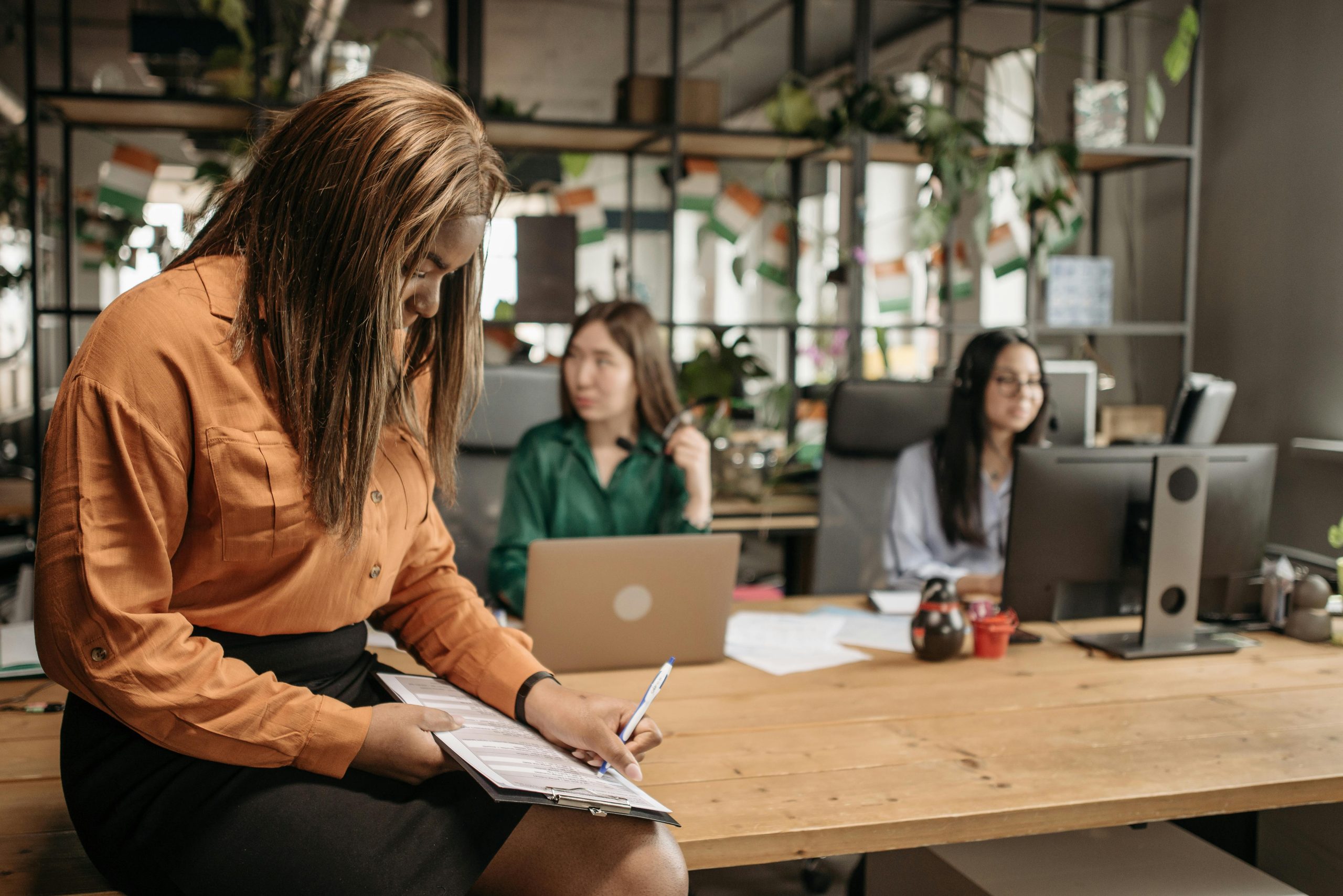 pexels-pavel-danilyuk-7658239 (1) A woman sitting on a conference table, writing notes on a clipboard, with two coworkers seated in the back.